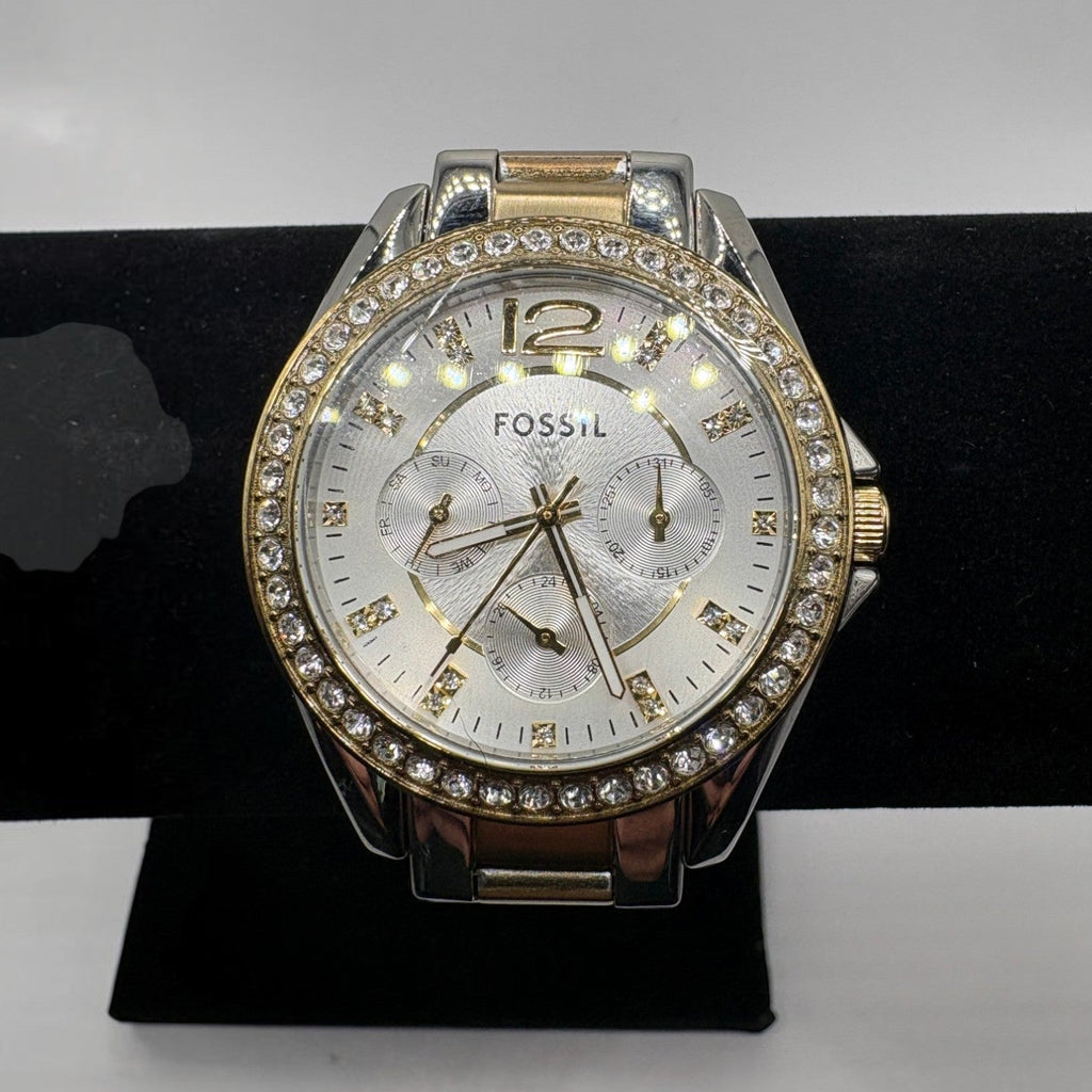 Fossil women's watch with a silver and rose gold-tone case, a crystal-studded bezel, and a white dial, displayed on a black stand.