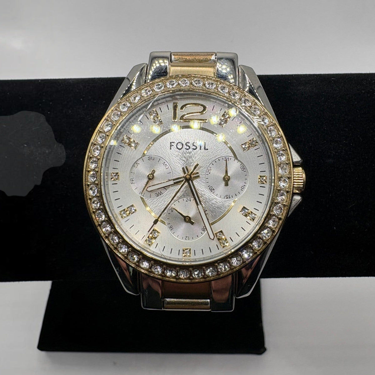Fossil women's watch with a silver and rose gold-tone case, a crystal-studded bezel, and a white dial, displayed on a black stand.